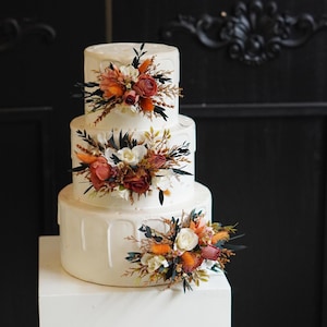 May include: A three-tiered white wedding cake decorated with cascading white frosting and orange, white, and brown floral arrangements.