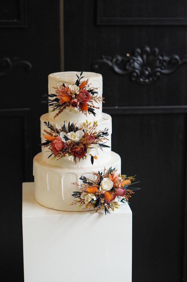May include: A three-tiered white wedding cake decorated with cascading white frosting and orange, white, and brown floral arrangements.