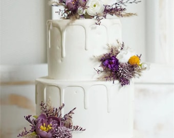 May include: A three-tiered white cake with a drip icing design. The cake is decorated with clusters of purple, white, and yellow flowers and dried grasses. The cake is likely for a wedding or special occasion.