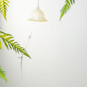 Pode incluir: Um ornamento em forma de sino creme pendura-se de um fio de barbante natural. Uma haste fina e transparente estende-se para baixo, com uma pequena conta de cristal. Frondes verdes de samambaia emolduram o ornamento contra um fundo branco.