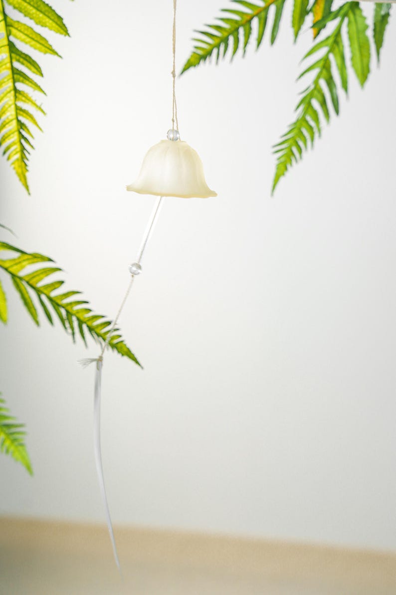 Pode incluir: Um ornamento em forma de sino creme pendura-se de um fio de barbante natural. Uma haste fina e transparente estende-se para baixo, com uma pequena conta de cristal. Frondes verdes de samambaia emolduram o ornamento contra um fundo branco.
