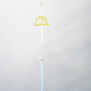Pode incluir: Um sino de vento pendurado com um topo em forma de sino amarelo. O sino tem uma haste transparente e fina e uma corda branca com pequenas contas transparentes. Uma etiqueta retangular pende na parte inferior.