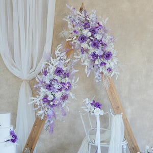 May include: A wedding ceremony setup featuring a white three-tiered cake adorned with purple flowers, a wooden arch decorated with purple and white floral arrangements, and a white chair with floral accents. Sheer white fabric drapes add an elegant touch.