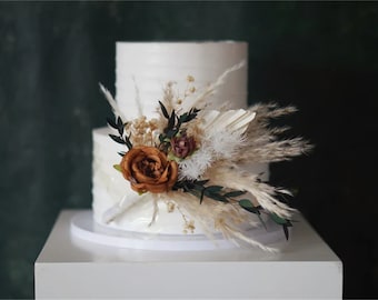 May include: Two-tiered white wedding cake decorated with a bouquet of dried pampas grass, brown roses, and greenery.