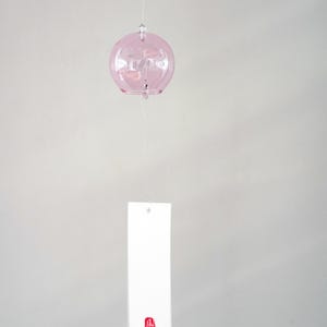 May include: A light pink, spherical glass wind chime suspended by a thin string. A rectangular white paper strip hangs from the bottom of the glass sphere, featuring a small, vertical red rectangular stamp with white Japanese characters at the base.