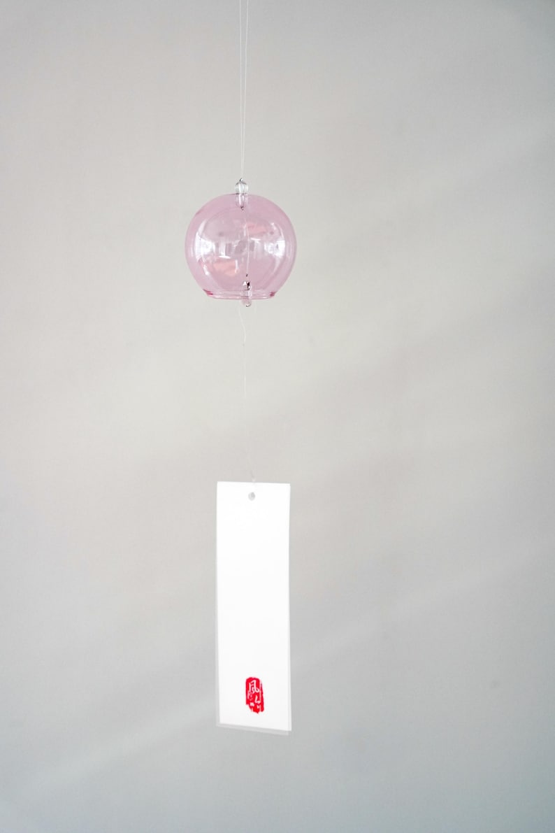 May include: A light pink, spherical glass wind chime suspended by a thin string. A rectangular white paper strip hangs from the bottom of the glass sphere, featuring a small, vertical red rectangular stamp with white Japanese characters at the base.
