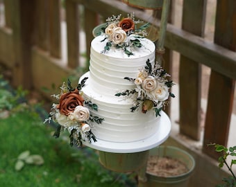 Op de afbeelding: Twee-laags witte bruidstaart versierd met bruine en witte bloemen. De taart staat op een witte taartstandaard en staat op een houten hek.