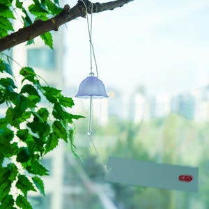 Pode incluir: Um delicado sino de vento lavanda com um design em forma de sino pendurado em um galho de &aacute;rvore. O sino tem uma haste longa e transparente e uma pequena conta redonda. Folhas verdes e uma paisagem urbana s&atilde;o vis&iacute;veis ao fundo.