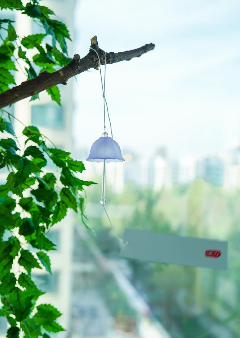 Pode incluir: Um delicado sino de vento lavanda com um design em forma de sino pendurado em um galho de &aacute;rvore. O sino tem uma haste longa e transparente e uma pequena conta redonda. Folhas verdes e uma paisagem urbana s&atilde;o vis&iacute;veis ao fundo.