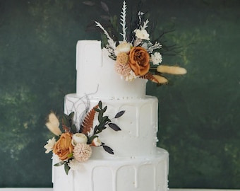 Op de afbeelding: Een drielaagse witte bruidstaart versierd met gedroogde bloemen en groen. De taart heeft een druppeleffect en is versierd met kleine witte stippen. De bloemen zijn een mix van bruin, wit en crème.