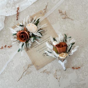 Puede incluir: Dos boutonnieres florales y un ramillete de muñeca a juego. Los boutonnieres y el ramillete presentan rosas color óxido, flores color crema y acentos plumosos. Están dispuestos sobre un papel vintage con texto y una tela de encaje blanco.