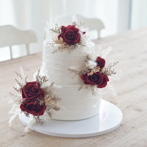 May include: A two-tiered white cake decorated with deep red roses and feathery, dried floral accents. The cake sits on a white cake stand, with a textured buttercream finish. The cake is a centerpiece for celebrations.