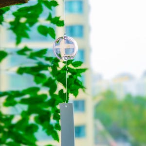 May include: A clear glass wind chime with a spherical bell hanging from a thin string. A rectangular, light gray paper strip is attached to the bottom of the glass sphere, featuring a small, red, rectangular stamp near the bottom edge.