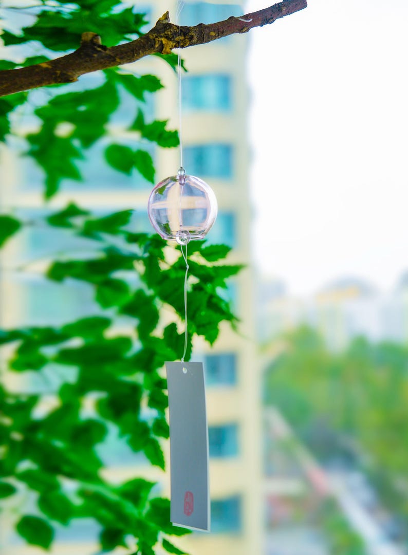 May include: A clear glass wind chime with a spherical bell hanging from a thin string. A rectangular, light gray paper strip is attached to the bottom of the glass sphere, featuring a small, red, rectangular stamp near the bottom edge.