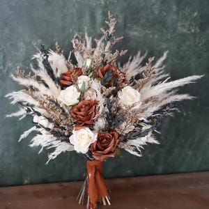 Può includere: Un bouquet di fiori con rose color ruggine e crema, soffice erba della pampa bianca, fogliame verde scuro e fiori secchi di riempimento marroni. Gli steli sono raccolti e legati insieme con un nastro di tessuto color ruggine alla base.