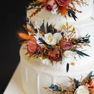 May include: A white wedding cake decorated with cascading floral arrangements. The flowers are a mix of white, orange, and burgundy.