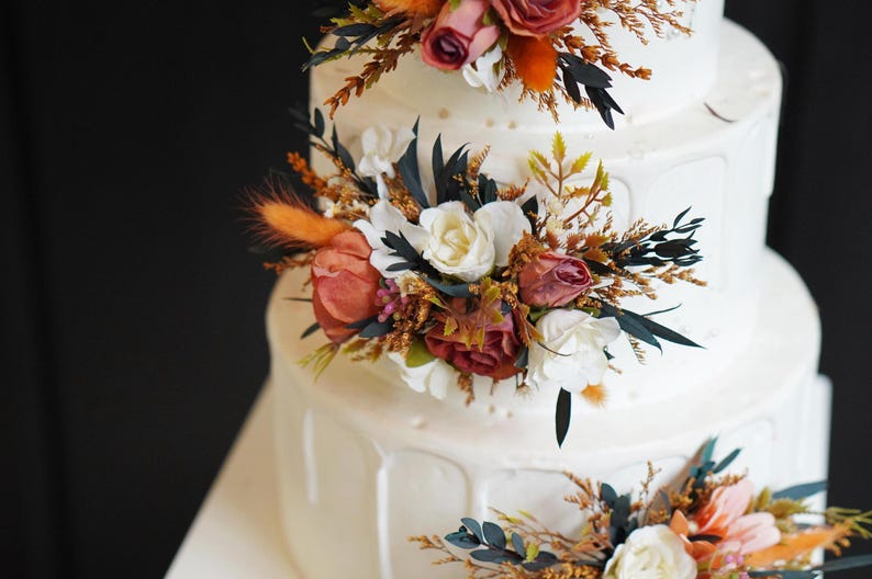 May include: A white wedding cake decorated with cascading floral arrangements. The flowers are a mix of white, orange, and burgundy.