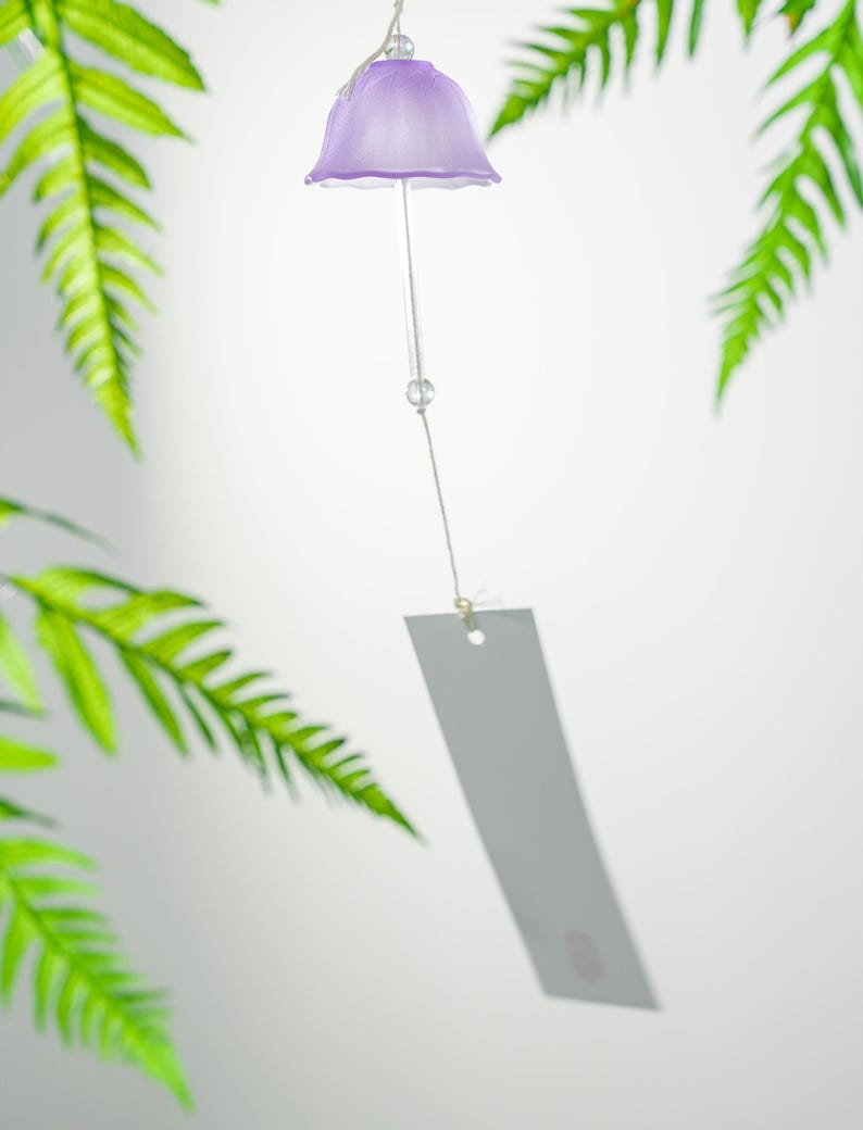 Pode incluir: Um delicado sino de vento cor de lavanda com um topo em forma de sino e uma etiqueta retangular pendurada. O sino pende de um fio fino, com folhas verdes de samambaia ao fundo. O sino &eacute; feito de vidro transl&uacute;cido.
