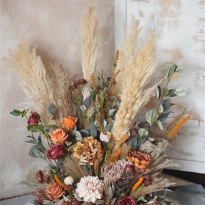 May include: A large floral arrangement with dried pampas grass, roses, and other flowers in shades of brown, orange, and pink. The arrangement is in a vase and is sitting on a gray surface.