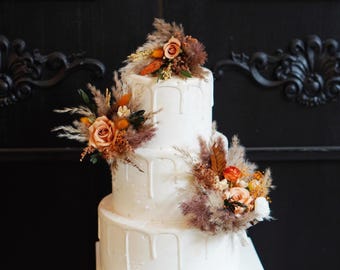Op de afbeelding: Een witte bruidstaart met drie lagen en een druipglazuurontwerp. De taart is versierd met arrangementen van gedroogde bloemen, waaronder rozen, pampasgras en andere texturele elementen in tinten perzik, oranje en bruin.