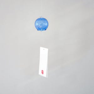 May include: A blue glass wind chime featuring a spherical bell shape suspended by a thin string. A rectangular white paper strip hangs from the bottom of the glass sphere, featuring a small red stamp mark near the bottom edge of the paper.