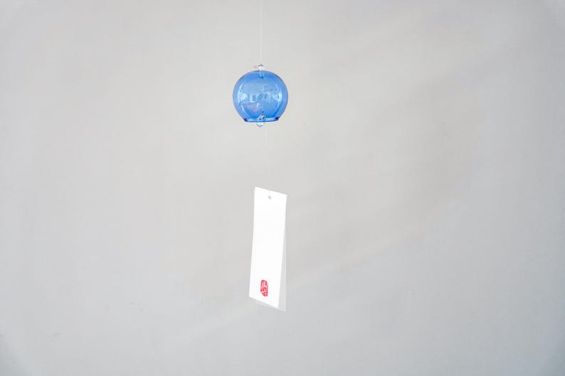 May include: A blue glass wind chime featuring a spherical bell shape suspended by a thin string. A rectangular white paper strip hangs from the bottom of the glass sphere, featuring a small red stamp mark near the bottom edge of the paper.
