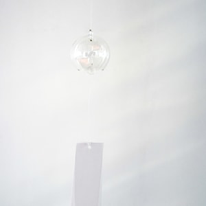 May include: A clear glass wind chime featuring a spherical bell suspended by a thin string. A rectangular white paper strip hangs below the bell, featuring a small red square stamp with the Japanese kanji character for wind printed in black.