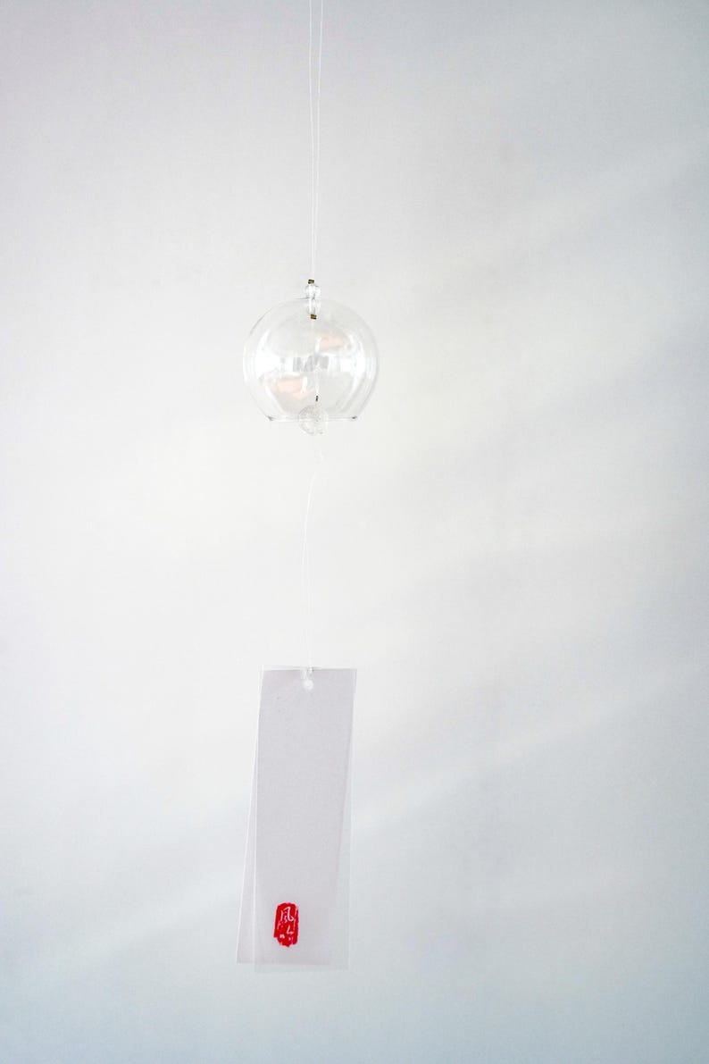 May include: A clear glass wind chime featuring a spherical bell suspended by a thin string. A rectangular white paper strip hangs below the bell, featuring a small red square stamp with the Japanese kanji character for wind printed in black.