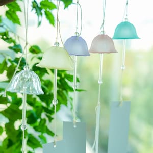 Pode incluir: Uma cole&ccedil;&atilde;o de sinos de vento em tons pastel pendurados em um galho de &aacute;rvore. Os sinos t&ecirc;m a parte superior em forma de sino em tons de transparente, amarelo, lavanda, rosa e verde-azulado. Cada sino tem um fio transparente com contas e uma etiqueta retangular.