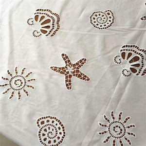 May include: White fabric with cut-out designs of sea creatures, including a starfish, sun, and seashells. The cut-outs reveal a brown background, creating a contrasting effect. The fabric is likely a tablecloth or decorative textile.