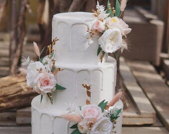 May include: A three-tiered white cake with a drip icing design. The cake is decorated with clusters of white and pink roses, green leaves, and gold accents. The cake is set on a wooden surface.