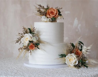 Op de afbeelding: Twee-laags witte bruidstaart versierd met gedroogde bloemen en pampasgras. De bloemen zijn in tinten perzik, crème en bruin.