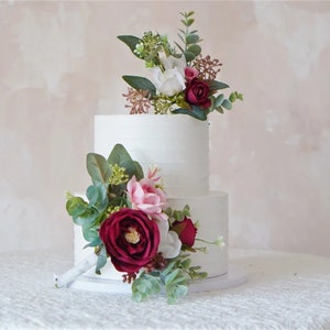 Può includere: Torta nuziale bianca a due piani decorata con un'arrangiamento floreale bordeaux e rosa. La torta è adornata con un bouquet di rose bordeaux, rose rosa e bianche, foglie di eucalipto e verde.
