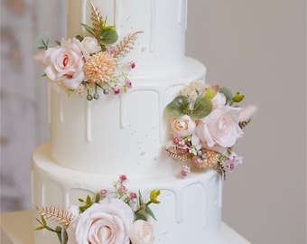 May include: A three-tiered white wedding cake with a drip icing design. The cake is decorated with pink and peach roses, greenery, and gold accents.