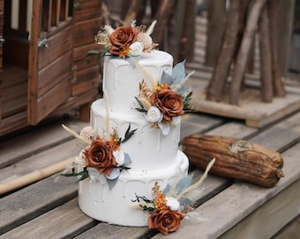 Op de afbeelding: Een witte taart met drie lagen en een druipontwerp, versierd met roestkleurige rozen, witte bloemen en gedroogd blad. De taart is versierd met accenten van blauwgrijze bladeren en staat op een houten oppervlak.