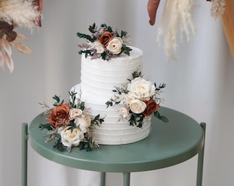 May include: A two-tier white cake with horizontal textured frosting, decorated with clusters of artificial flowers in shades of rust orange, cream, and white, accented with dark green foliage and small dried filler plants.
