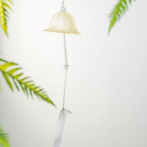 Pode incluir: Um sino de vento creme com um topo em forma de sino e uma haste transparente pendurada. O sino est&aacute; pendurado em uma corda, com uma pequena conta transparente. Folhas verdes de samambaia no fundo.