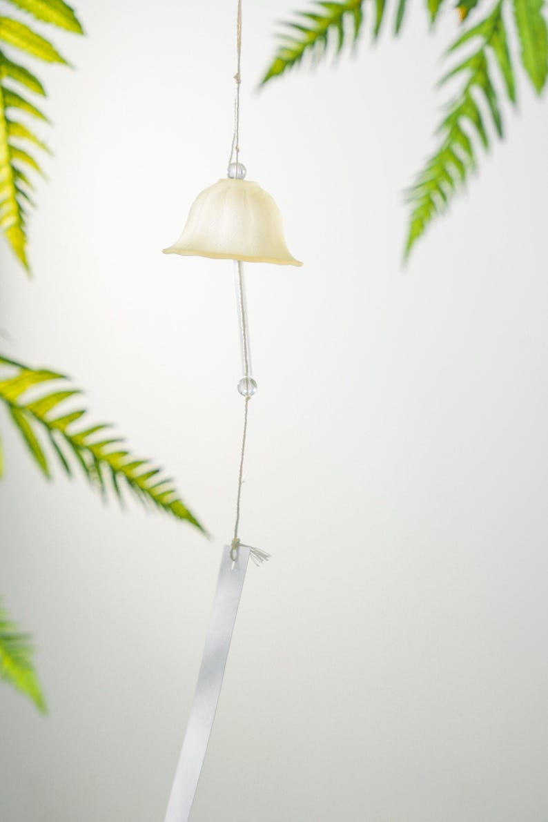 Pode incluir: Um sino de vento creme com um topo em forma de sino e uma haste transparente pendurada. O sino est&aacute; pendurado em uma corda, com uma pequena conta transparente. Folhas verdes de samambaia no fundo.