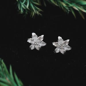 May include: A pair of silver flower stud earrings. Each earring has a detailed floral design with multiple petals. The earrings are set against a black background, with green foliage in the corners.