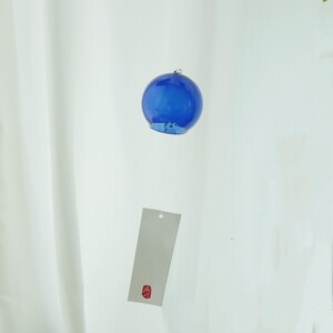 May include: A spherical blue glass wind chime featuring a smooth, translucent surface and a small metal loop at the top. A rectangular white paper strip hangs below the glass sphere, displaying a small red square stamp with black Japanese text.
