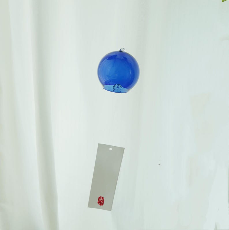 May include: A spherical blue glass wind chime featuring a smooth, translucent surface and a small metal loop at the top. A rectangular white paper strip hangs below the glass sphere, displaying a small red square stamp with black Japanese text.