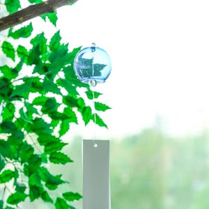 May include: A light blue spherical glass wind chime hanging from a branch. The chime features a clear glass clapper inside the sphere and a long, rectangular white paper strip attached by a thin string, marked with a small red Japanese character.
