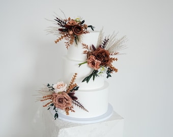 Op de afbeelding: Een witte taart met drie lagen versierd met bloemstukken. De bloemen zijn in tinten bruin, perzik en wit, met groen en gedroogde grassen. De taart staat op een witte sokkel tegen een witte achtergrond.
