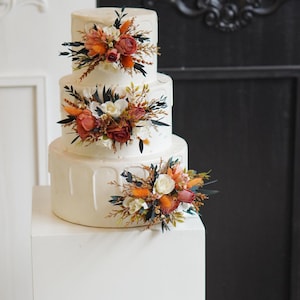 May include: A three-tiered white wedding cake decorated with cascading white icing and floral arrangements. The flowers are a mix of white, orange, and red.