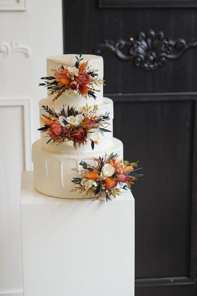 May include: A three-tiered white wedding cake decorated with cascading white icing and floral arrangements. The flowers are a mix of white, orange, and red.