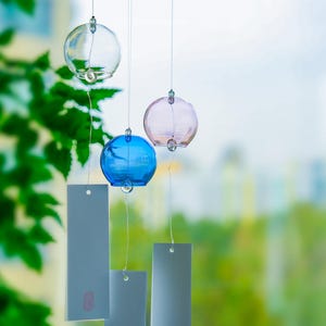 May include: A wind chime featuring three spherical glass ornaments in clear, light pink, and blue colors. Each glass sphere is suspended by a thin string and attached to a rectangular white paper tag that hangs vertically below the glass.