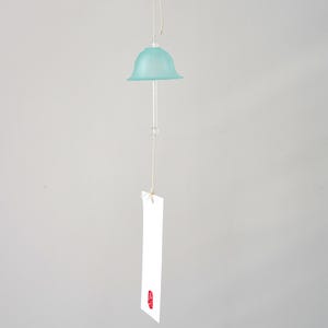 Pode incluir: Um sino de vento suspenso com um topo em forma de sino azul claro e uma haste de vidro transparente. Uma etiqueta retangular branca com um selo vermelho pende por baixo. O sino &eacute; suspenso por um fio fino e bege contra um fundo cinza.