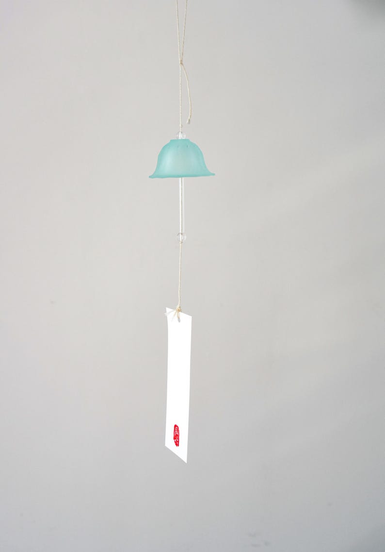 Pode incluir: Um sino de vento suspenso com um topo em forma de sino azul claro e uma haste de vidro transparente. Uma etiqueta retangular branca com um selo vermelho pende por baixo. O sino &eacute; suspenso por um fio fino e bege contra um fundo cinza.
