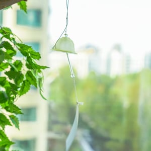 Pode incluir: Um pequeno sino de vento esbranqui&ccedil;ado pende de um fio fino, com um fundo desfocado de folhagem verde e edif&iacute;cios. O sino tem um topo em forma de sino e uma pe&ccedil;a retangular longa e fina pendurada abaixo. Folhas verdes vis&iacute;veis em primeiro plano.