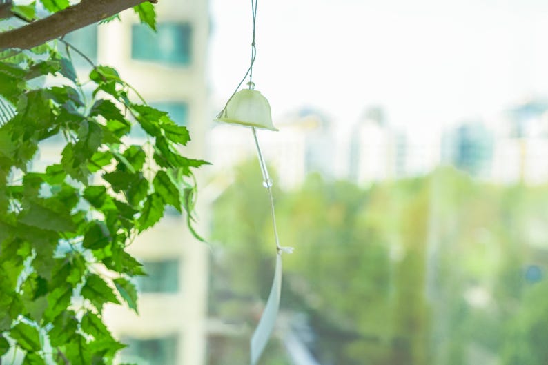 Pode incluir: Um pequeno sino de vento esbranqui&ccedil;ado pende de um fio fino, com um fundo desfocado de folhagem verde e edif&iacute;cios. O sino tem um topo em forma de sino e uma pe&ccedil;a retangular longa e fina pendurada abaixo. Folhas verdes vis&iacute;veis em primeiro plano.